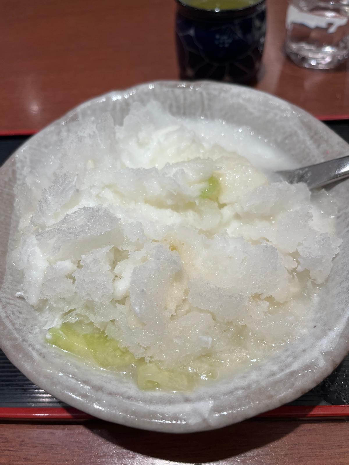白い器に入ったかき氷。粗く削られた氷が盛り付けられ、底にはキュウリが少量見える。背景には飲み物が置かれている。