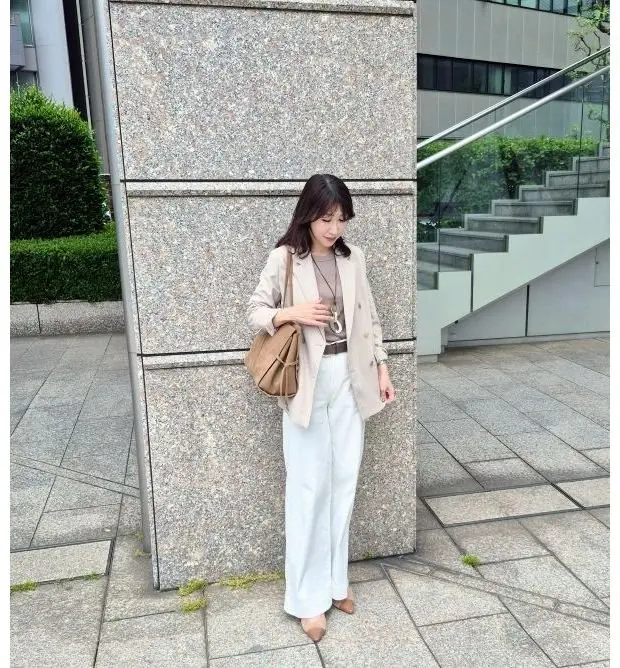 ベージュのジャケットと白いパンツを着た女性が、石造りの柱に寄りかかり、右手にベージュのハンドバッグを下げている。背景には手すり付きの階段と低木が見える。