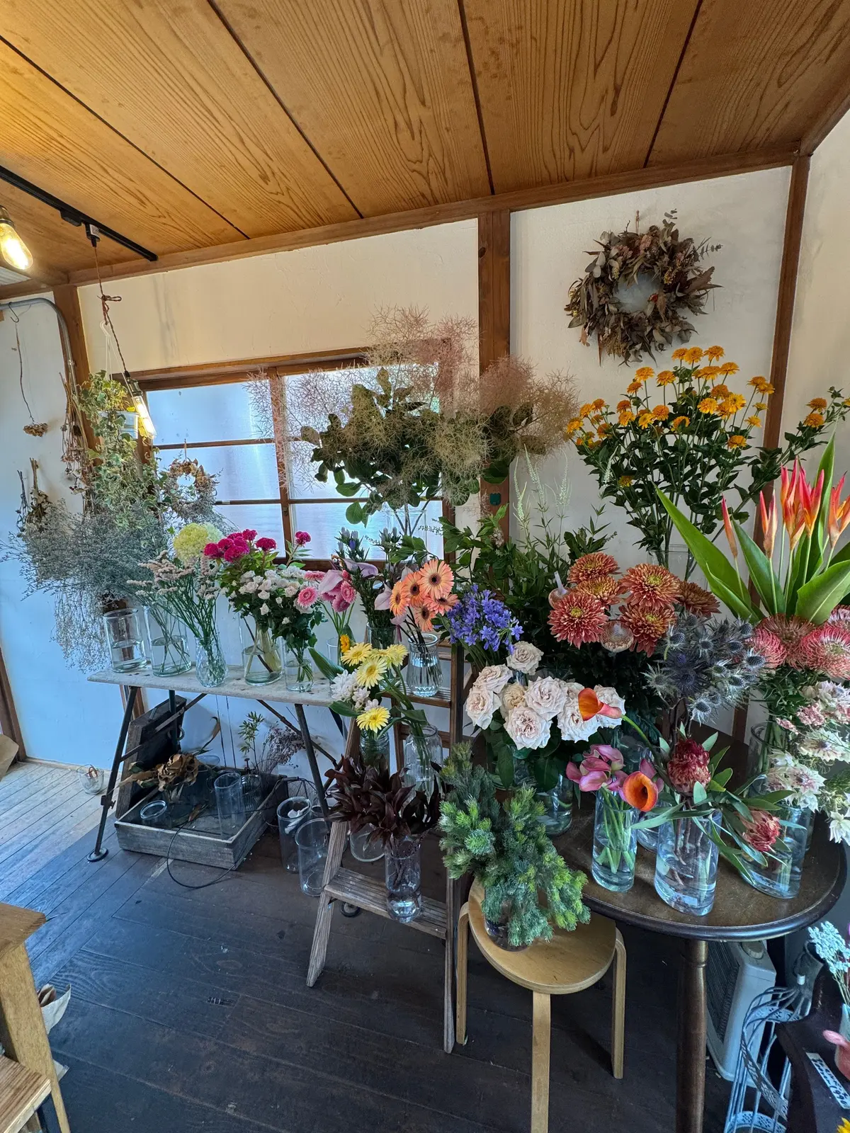 木造の天井と白い壁に囲まれた花屋の店内。様々な種類の花束や切り花が、複数のテーブルや棚に飾られている。ドライフラワーや緑の植物も多く、自然な雰囲気が感じられる。壁には花環が飾られている。