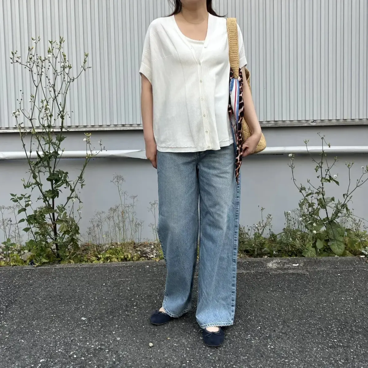 ベージュの半袖シャツとデニムパンツを着た女性が、ストライプ模様のショルダーバッグを肩にかけ、コンクリート地に立っています。背景には小さな白い花が咲く低木が並んでいます。