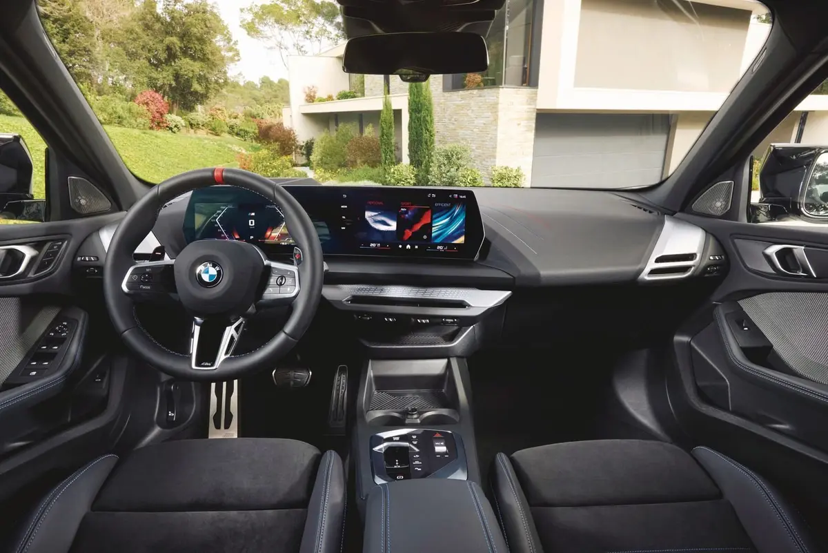 BMW i4の運転席内部。黒を基調とした内装に青色のアクセントが施され、中央に大型タッチスクリーン、右下にエアコン操作パネル、左には運転席用スイッチパネルが見える。ステアリングホイールにはBMWのロゴが配置。