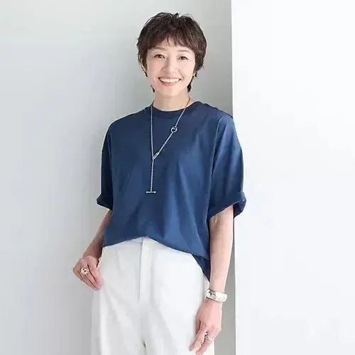 紺色のTシャツに白いパンツを着用した女性。短くカットした茶色の髪で、笑顔を見せている。シルバーのロングネックレスとリング、腕時計を身に着けている。背景は白。