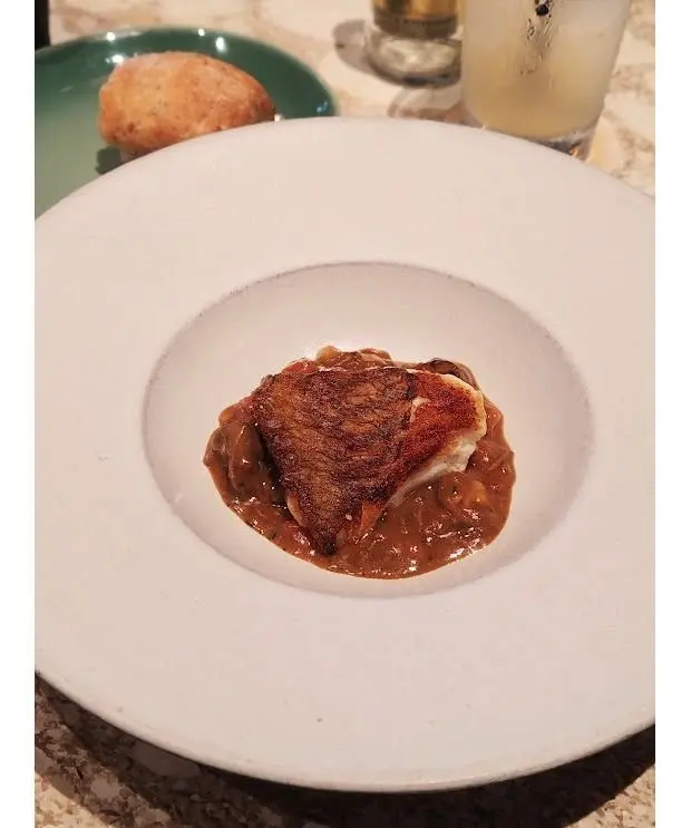 白磁の皿に盛り付けられた、表面に焼き色がついた魚の切り身。周りには茶色のソースと、薄切りにした玉ねぎが添えられている。背景には緑色の皿とグラスが見える。