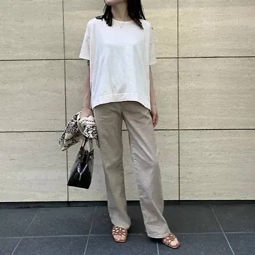 白の半袖シャツとベージュのワイドパンツを着た女性が、壁に寄りかかりながら立っている。左手には模様のある布と黒のハンドバッグを持っている。足元はサンダル。