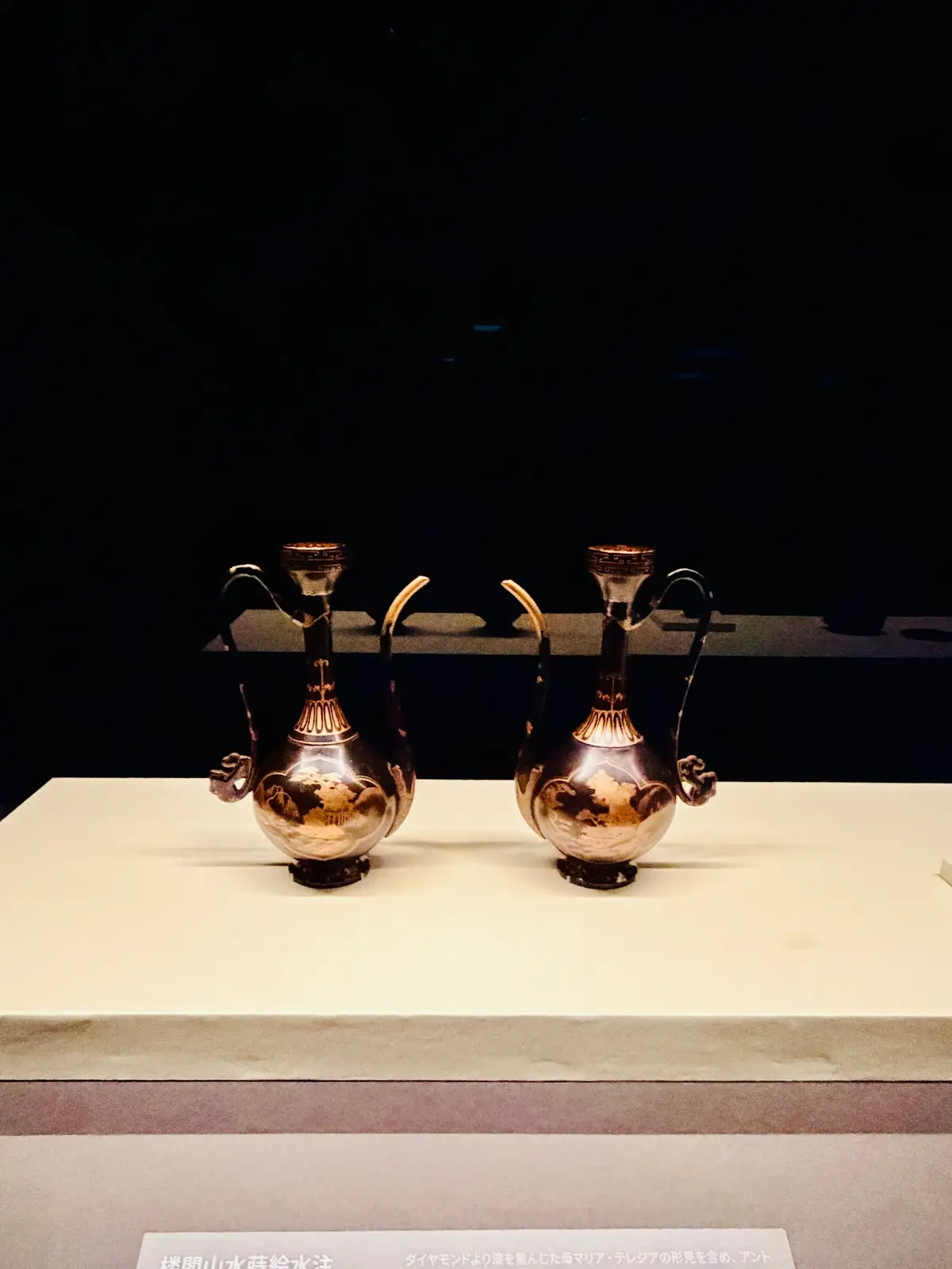 博物館の展示ケースに置かれた、金色の古代ギリシャの水差し2つ。胴体には模様が施され、それぞれ2つの取っ手と注ぎ口を持つ。