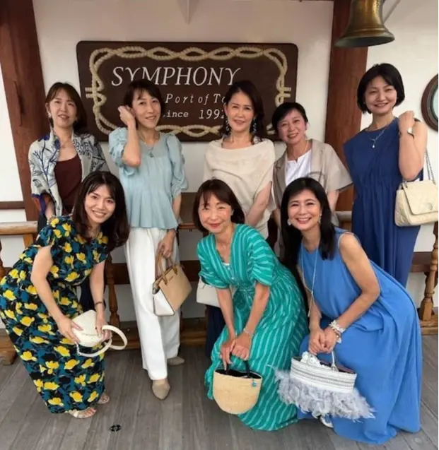 木製の甲板に、鮮やかな色の服を着た9人の女性が笑顔で写っている。背景には「SYMPHONY Port of Texas since 1992」と書かれた看板と、大きな船のベルが見える。