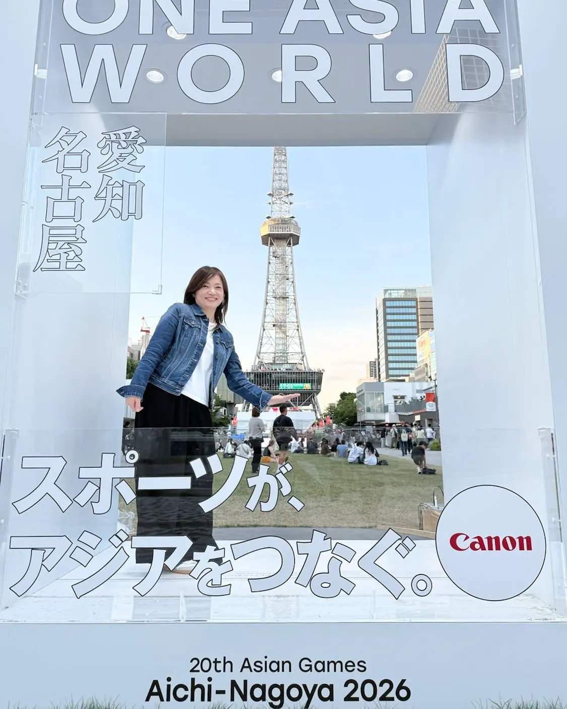 白いフレームの前で女性がポーズをとっている。背景には名古屋テレビ塔が見える。フレームには「ONE ASIA WORLD」の文字と、2026年愛知・名古屋アジア競技大会のロゴ、Canonのロゴが配置されている。