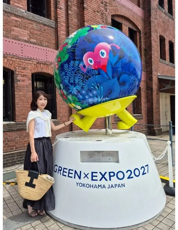 横浜で開催された「GREEN×EXPO 2027」のモニュメントの前で、女性がポーズを取っている。モニュメントは白地に青と緑の地球儀と鳥のイラストが描かれ、黄色い台座に乗っている。女性は白いトップスに茶色のスカート、麦わら帽子を着用。