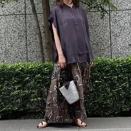 濃い紫色のボタンダウンシャツに、花柄のロングスカートを合わせた女性。黒いサンダルを履き、シルバーの小さなバッグを持つ。背景には、緑の低木とグレーの建物がある。