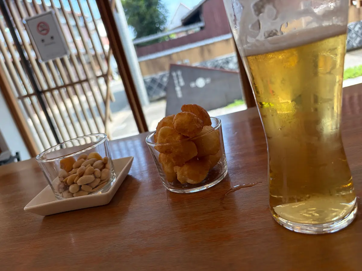 木製のテーブルに置かれた、ビール、ピーナッツ、ポテトチップスの盛り合わせ。ビールは泡立ち、グラスの右側に位置する。