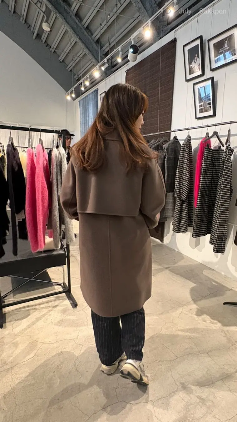 グレーのオーバーサイズコートを着た女性が、衣料品店で後ろを向いて立っている。背景には、さまざまな服やアクセサリーが並んだラックが見える。天井にはスポットライトが設置されている。