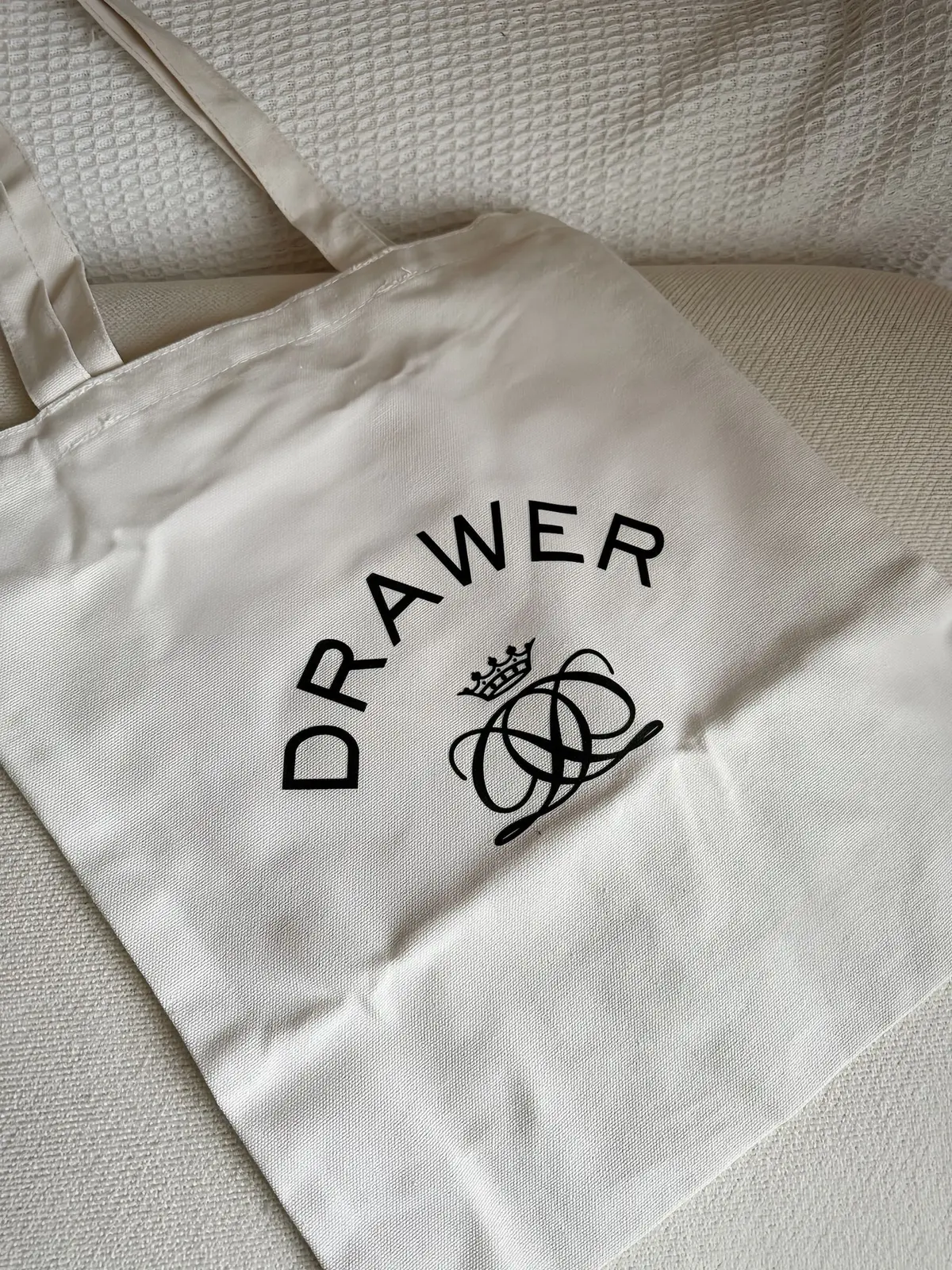 白いキャンバストートバッグ。中央に黒色の「DRAWER」の文字が弧を描くように配置され、その下に王冠と絡み合ったリボンをモチーフにしたロゴマークが印刷されている。