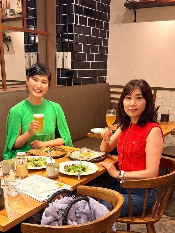 レストランで食事を楽しむ2人の女性。左の女性は緑色のトップスにビール、右の女性は赤いトップスにワイングラスを持っている。テーブルにはサラダや料理が並び、背景には黒いタイルが目立つ。