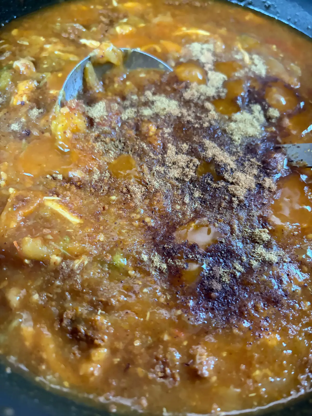 黒い鍋の中で、鶏肉と野菜が煮込まれた濃い茶色のスープ。中央にはスパイスが振りかけられ、金属製のヘラがスープをかき混ぜている。