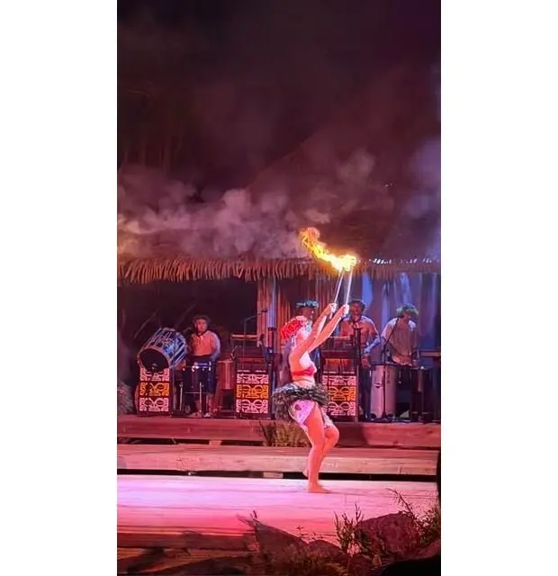 夜のステージで、女性が炎のマリンバを高く掲げ、踊っている。背景には、太鼓を叩く男性や、楽器を演奏する男性がいる。藁葺き屋根の小屋と、煙が立ち込める様子も見える。