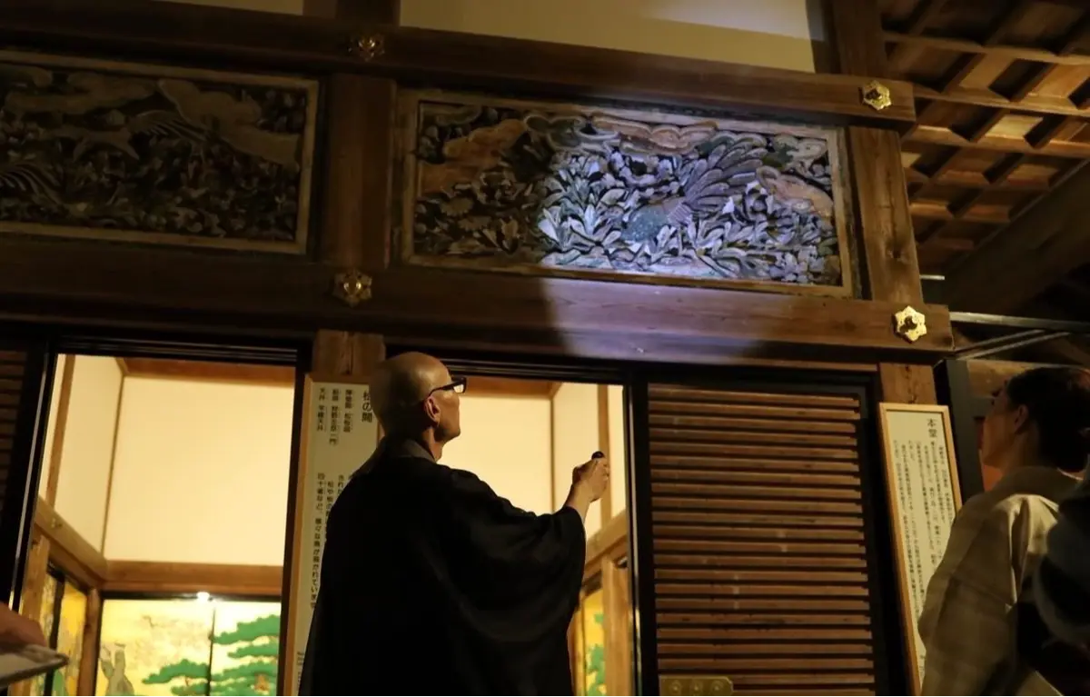 寺の入り口で、黒い法衣を着た男性が説明をしている様子。男性は片手で何かを指し示し、もう一方の手には懐中電灯を持っている。背景には精緻な彫刻が施された木製の装飾がある。