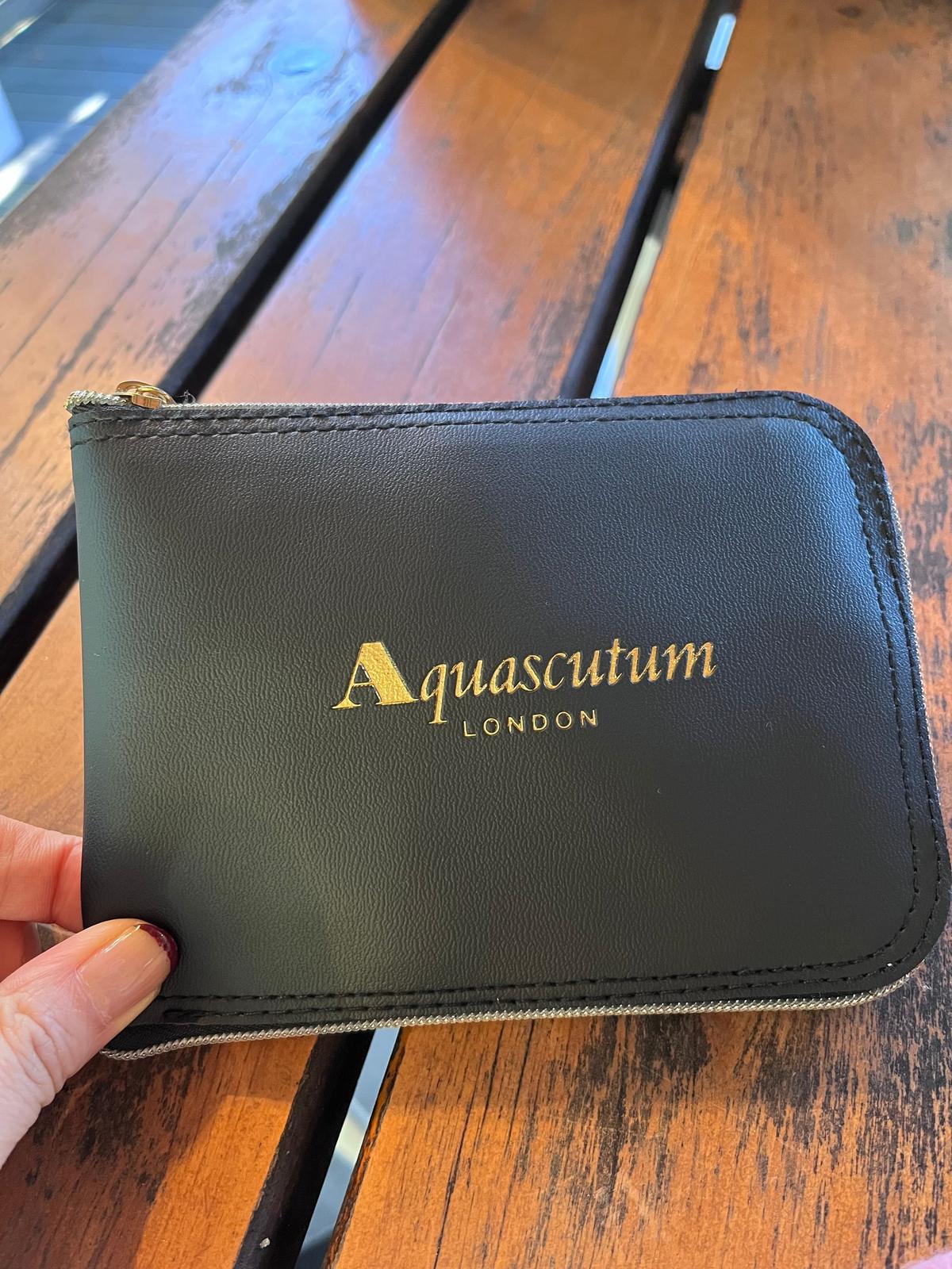 黒いレザーのカードケース。前面中央に金色の「Aquascutum LONDON」のロゴが配置されている。ジッパー付きで、手で保持されている。