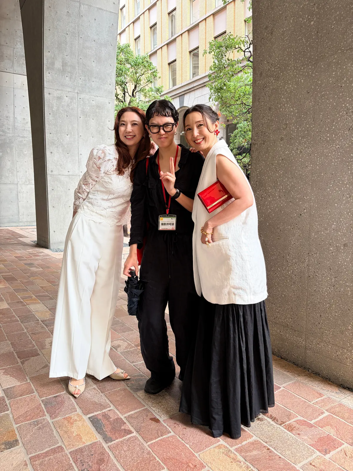 3人の女性が笑顔で並んで写っている写真。左は白いパンツスーツ、右は白いベストと黒いロングスカート、中央は黒いパンツとトップス。背景にはコンクリートの建物と緑の木々が見える。