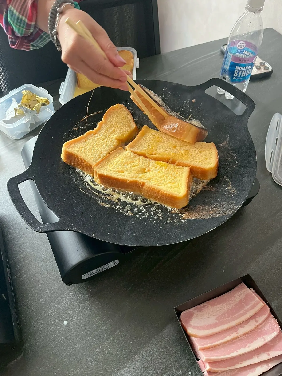 お箸でパンの片面をひっくり返す、フレンチトーストを作っている。焦げ目がついたパンが4枚並ぶ。テーブルには水のボトルやベーコンの入った箱がある。