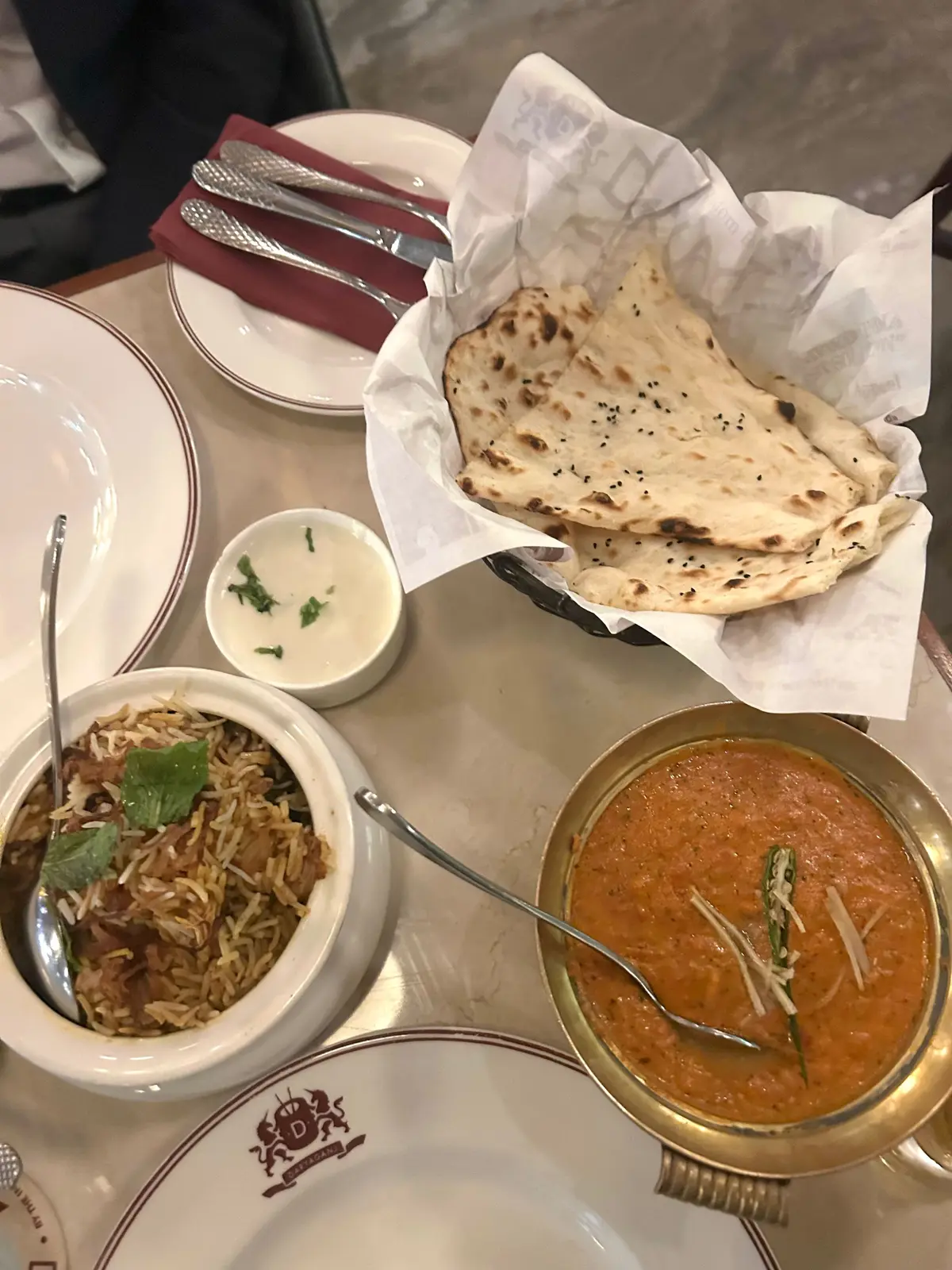 テーブルに置かれたインド料理。白米のピラフ、ナン、カレーが盛り付けられている。小皿にはヨーグルト、カトラリーが添えられている。