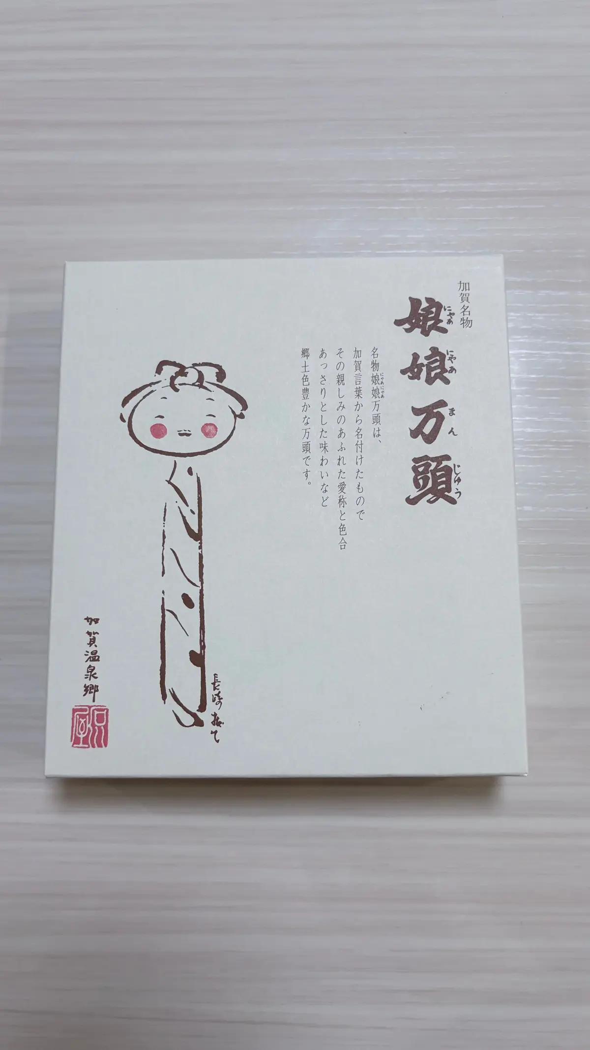 白い箱に、黒い線で描かれた丸い顔の人物イラストが描かれている。箱には日本語で「眼鏡万頭」と記載され、その下に説明文、右下に赤いロゴマークがある。
