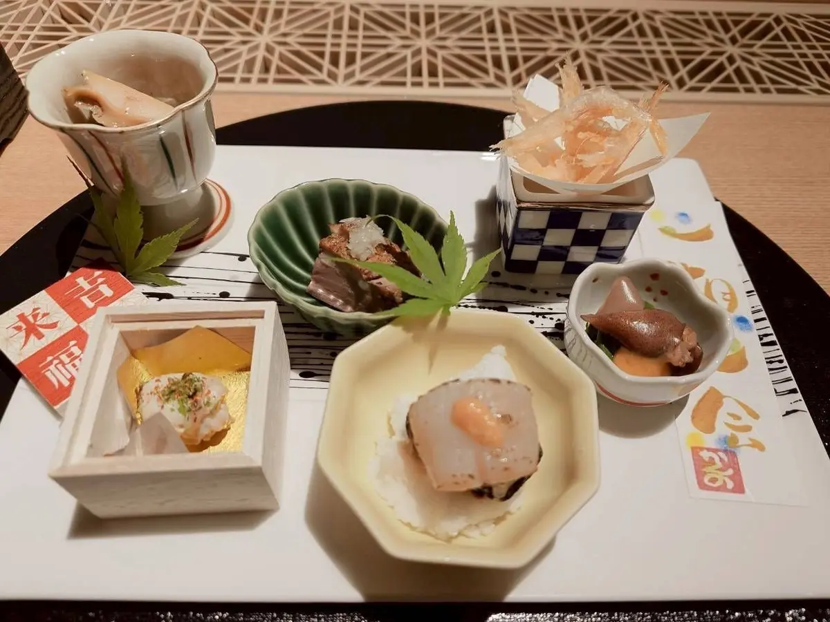 白いテーブルに、さまざまな料理が精巧に盛り付けられています。八角形の器に卵と白身魚、木製の箱に黄色の料理、緑の椀に肉と野菜、小さな白い器に魚の刺身などがあります。