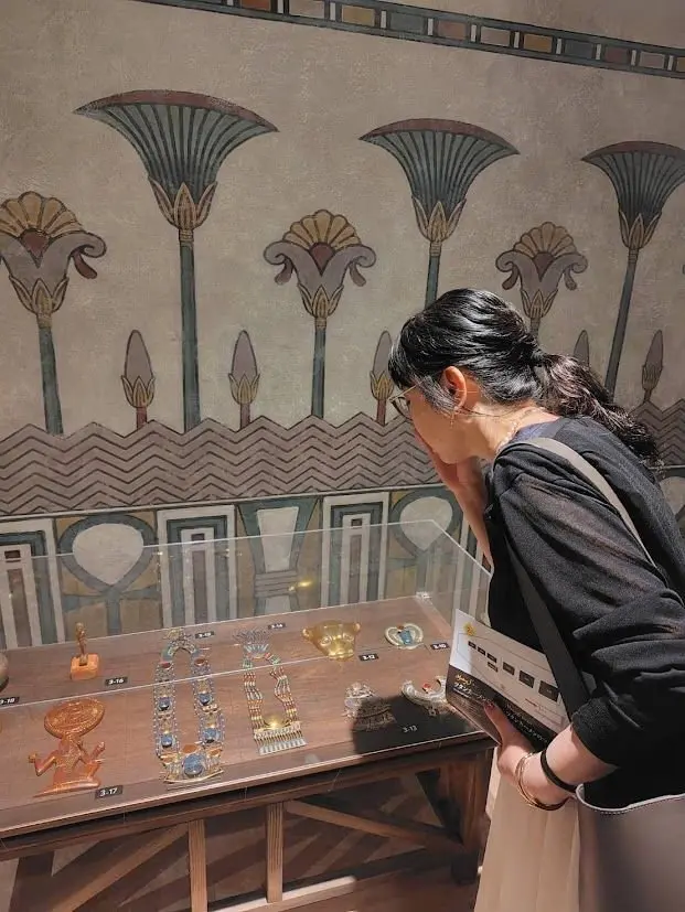 博物館で展示ケースを覗き込む女性。背景には古代エジプト風の壁画が描かれ、ケース内には小さな模型や装飾品が並んでいる。