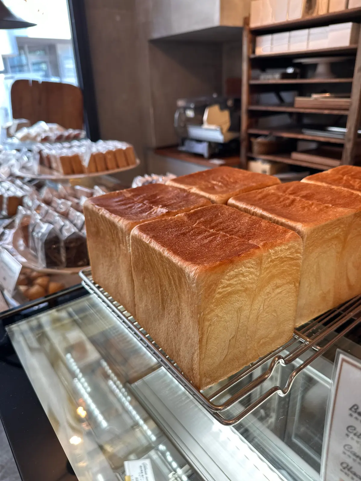 ショーケースに並んだ、焼き色のついた食パン。四角い形をしており、断面からふっくらとした内部が見える。背景には、他のパンや棚がある。