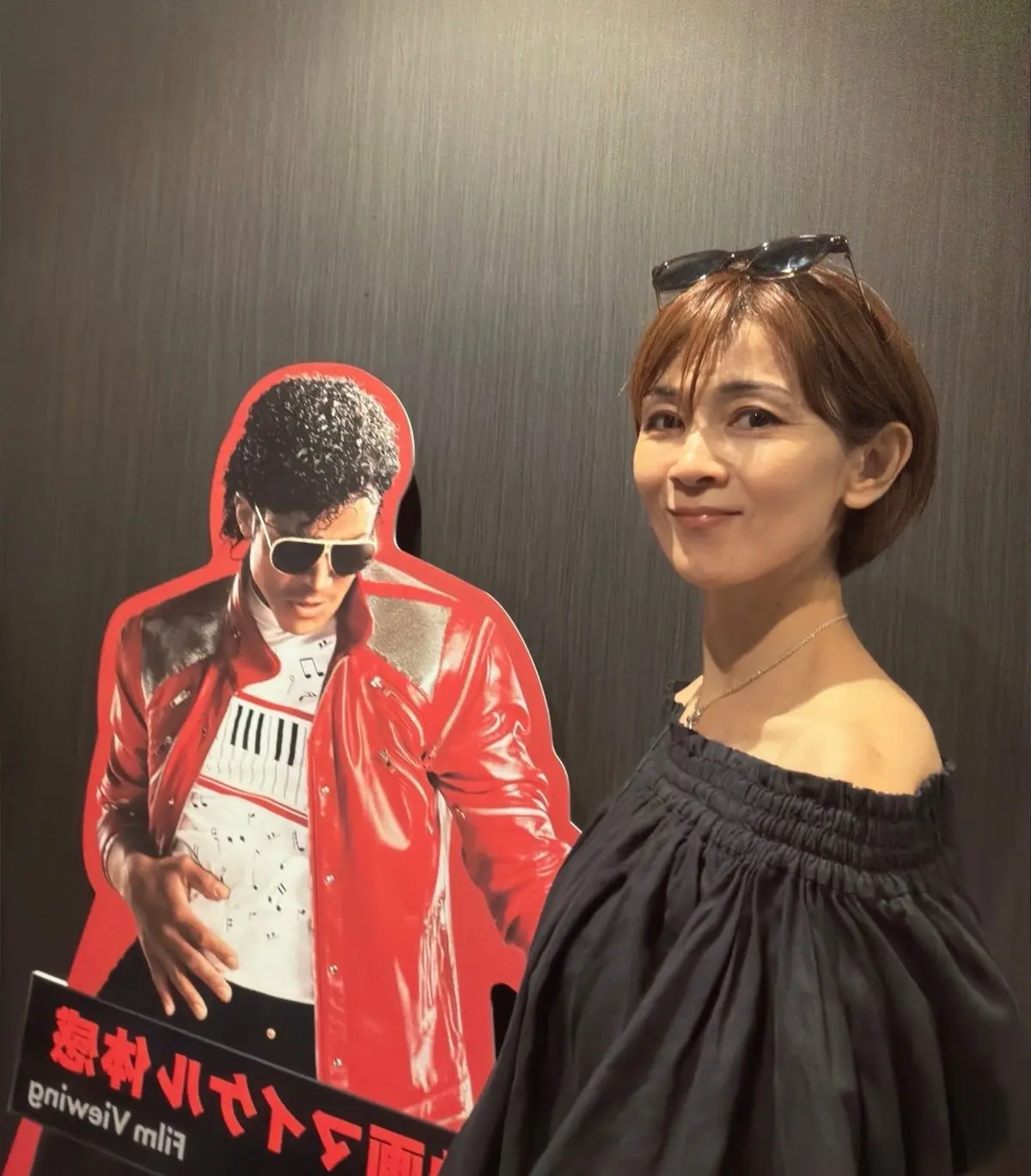 笑顔の女性が、マイケル・ジャクソンの等身大パネルの横に立っている写真。女性は黒いガウンを着用し、茶色のショートヘア。パネルは赤と黒で、マイケル・ジャクソンがピアノを弾いている姿が描かれている。