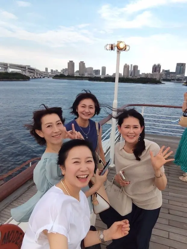 水上遊覧船のデッキで、5人の女性が笑顔で写っている。手前に白いTシャツを着た女性がおり、その他はそれぞれ青、ベージュ、水色の服を着ている。背景には橋と高層ビル群、水面が広がり、晴天の空が広がっている。