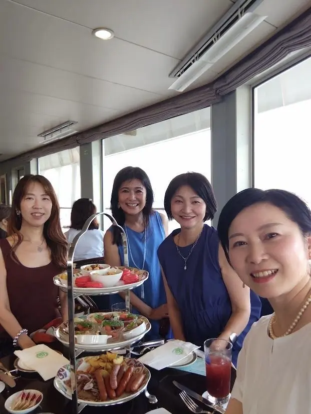 4人の女性が、三段重ねの豪華なアフタヌーンティーセットを囲んで笑顔。窓際のテーブルで、軽食や飲み物が並び、明るい雰囲気。