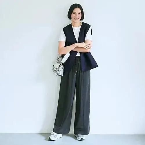 女性が白い壁に寄りかかり、笑顔でポーズしている。白のTシャツに紺のベストを着て、紺のワイドパンツを履き、右手に銀色のクラッチバッグを持っている。足元には白いスニーカーが見える。