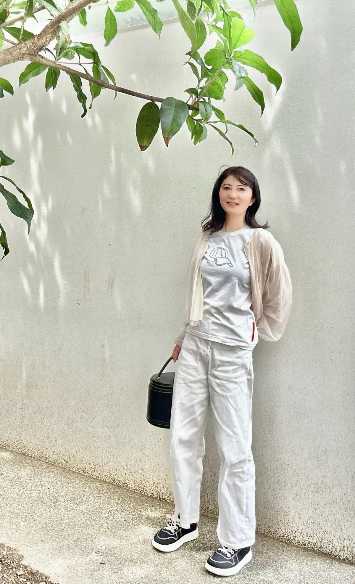 女性が白い壁に寄りかかり、軽い色の服を着ています。手には小さな黒いかごバッグを持ち、明るい日差しが葉の影を投げかけています。