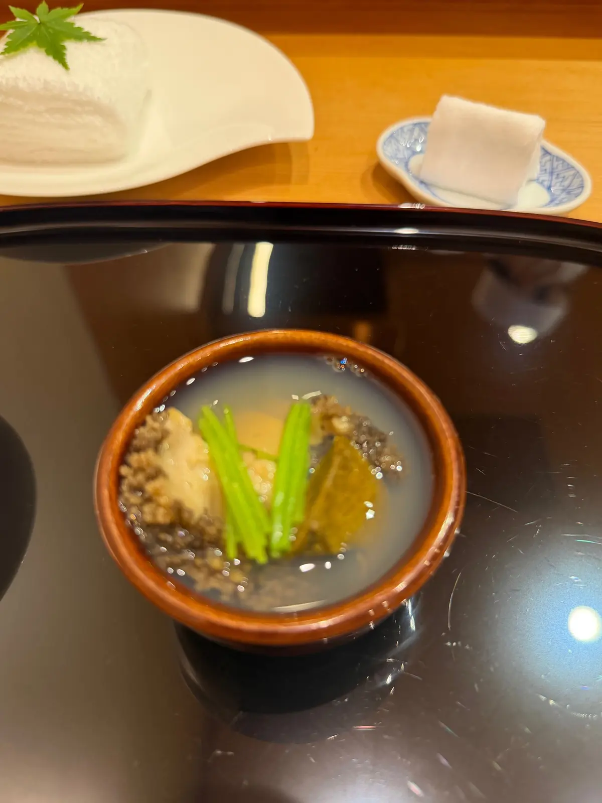 茶色の陶器鉢に入った透明な汁物。緑色の野菜が数本、茶色い具材と共に見える。背景には白いご飯と、模様のついた小さな皿が置かれている。