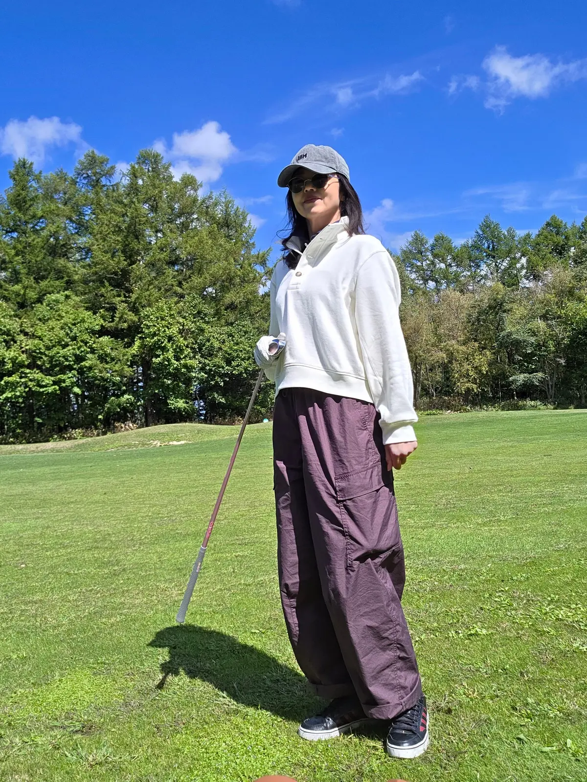 ゴルフコースに立つ女性。白いプルオーバー、紫色のゆったりしたパンツ、黒いキャップを着用。左手にゴルフクラブを持ち、向かい側にゴルフボールが見える。背景には緑の芝生と木々、青空が広がる。