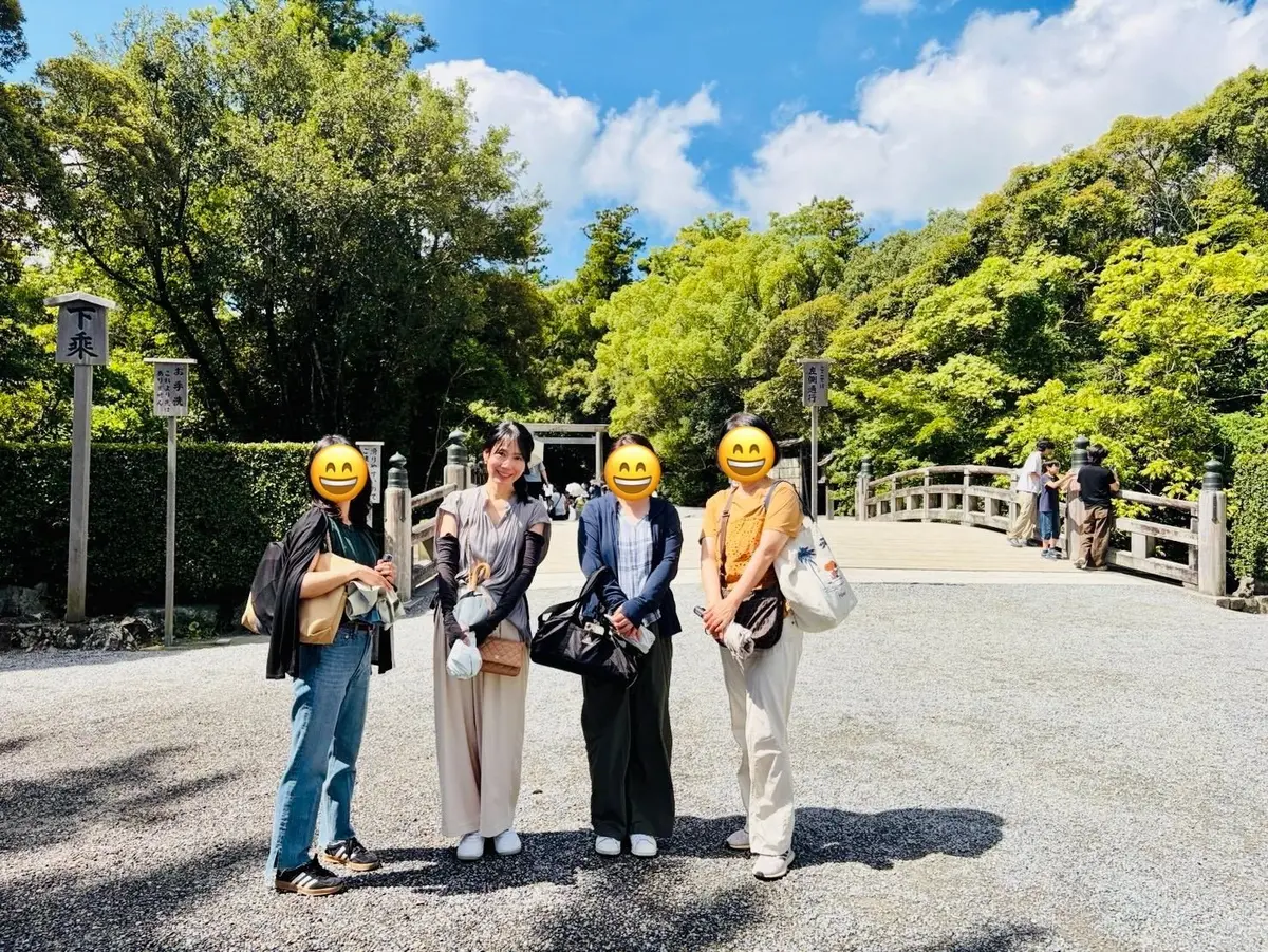 晴天の下、4人の女性が並んで立っている。左から、デニムの女性、ベージュのワンピースの女性、紺色のジャケットの女性、黄色のトップスとベージュのパンツの女性。背景には緑豊かな木々と、石造りの橋が見える。