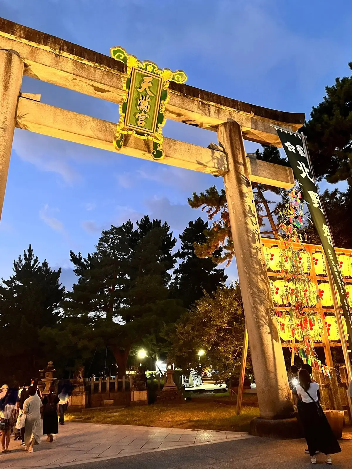 夕暮れ時の北野天満宮。鳥居が正面にあり、緑色の額には「天満宮」の文字。右側には提灯が並び、人々が参拝している。