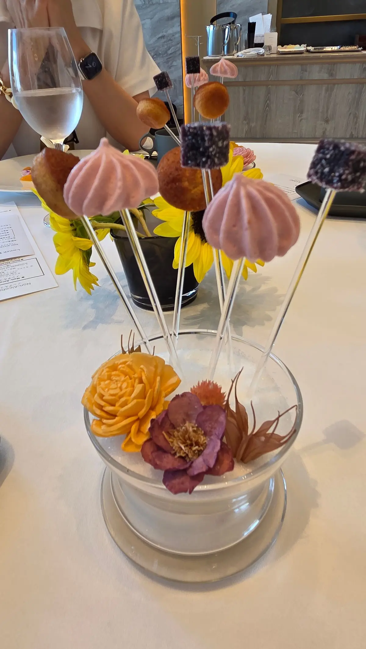 透明な器に、ピンクや茶色の可愛らしいお菓子が串に刺されて飾られている。黄色い花や紫色の花も添えられ、上品なテーブルコーディネート。