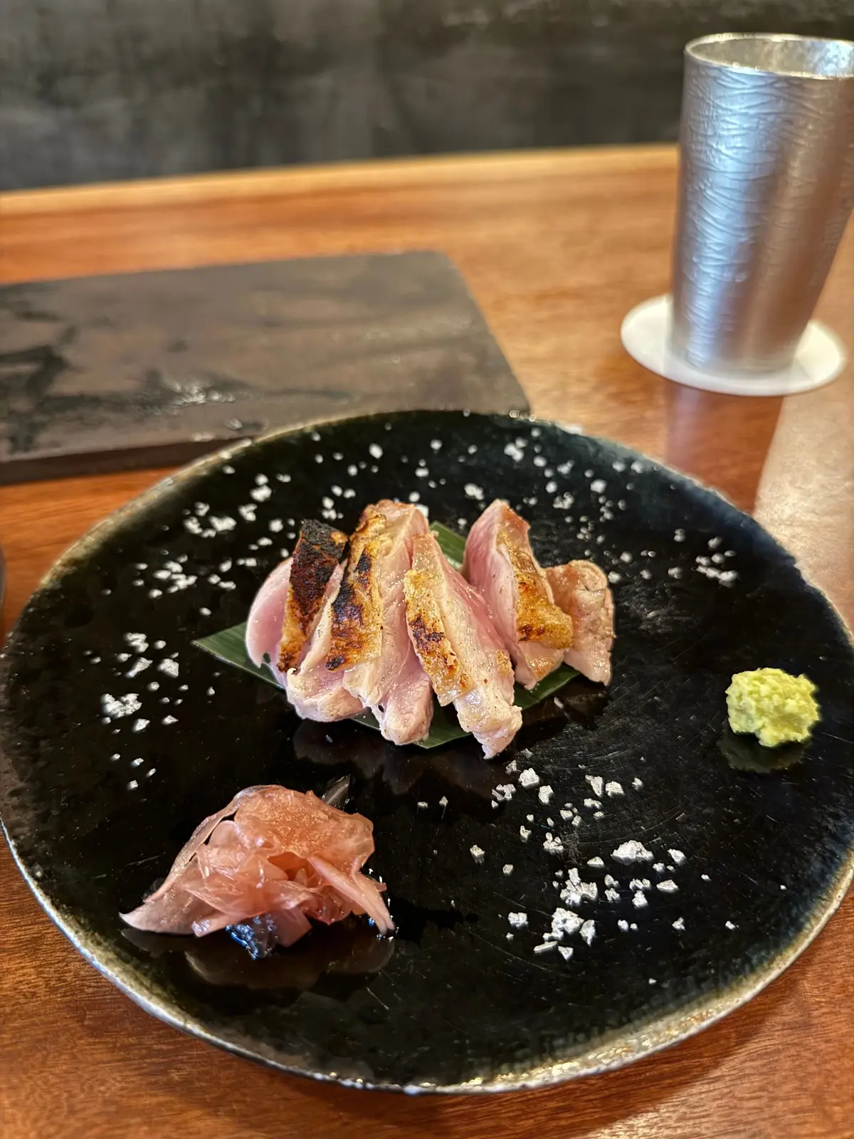 大阪・福島の人気焼き鳥店「焼き鳥 小松」でいただいた焼き鳥コース。黒い皿に盛り付けられた、照り焼き風味の鴨肉とピクルス、わさび。鴨肉は3切れ、緑色の葉の上に置かれ、皿全体には白い粉が散らされている。
