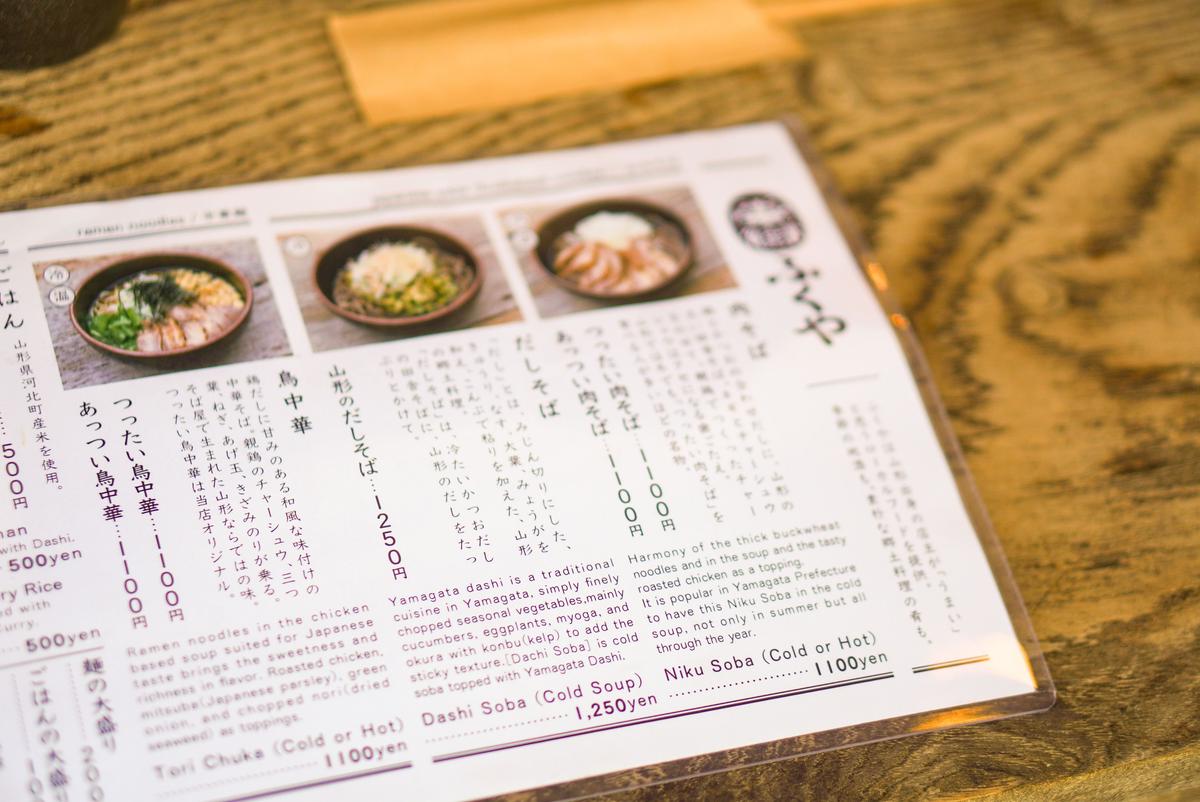 手書き風の日本語メニュー。ラーメン、そば、ご飯などの料理名と値段が記載。右ページには、山形だしを使った料理の説明と写真が掲載されている。全体的に茶色と白を基調とした落ち着いた色合い。
