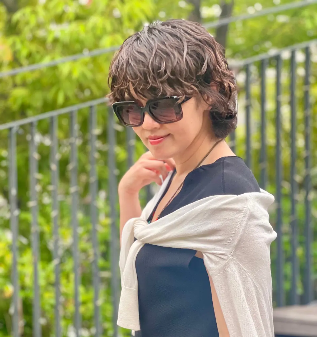 短い巻き毛の女性が、黒いサングラスをかけ、白いショールと紺色のドレスを着て、屋外で微笑んでいます。背景には緑の植物とフェンスが見えます。