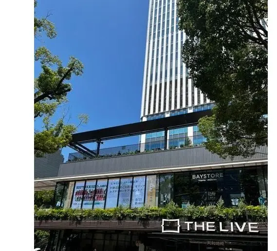 高層ビルと商業施設「THE LIVE」のファサード。青空を背景に、近代的なデザインの建物が写る。手前には歩行者がおり、明るい日差しが照りつけている。
