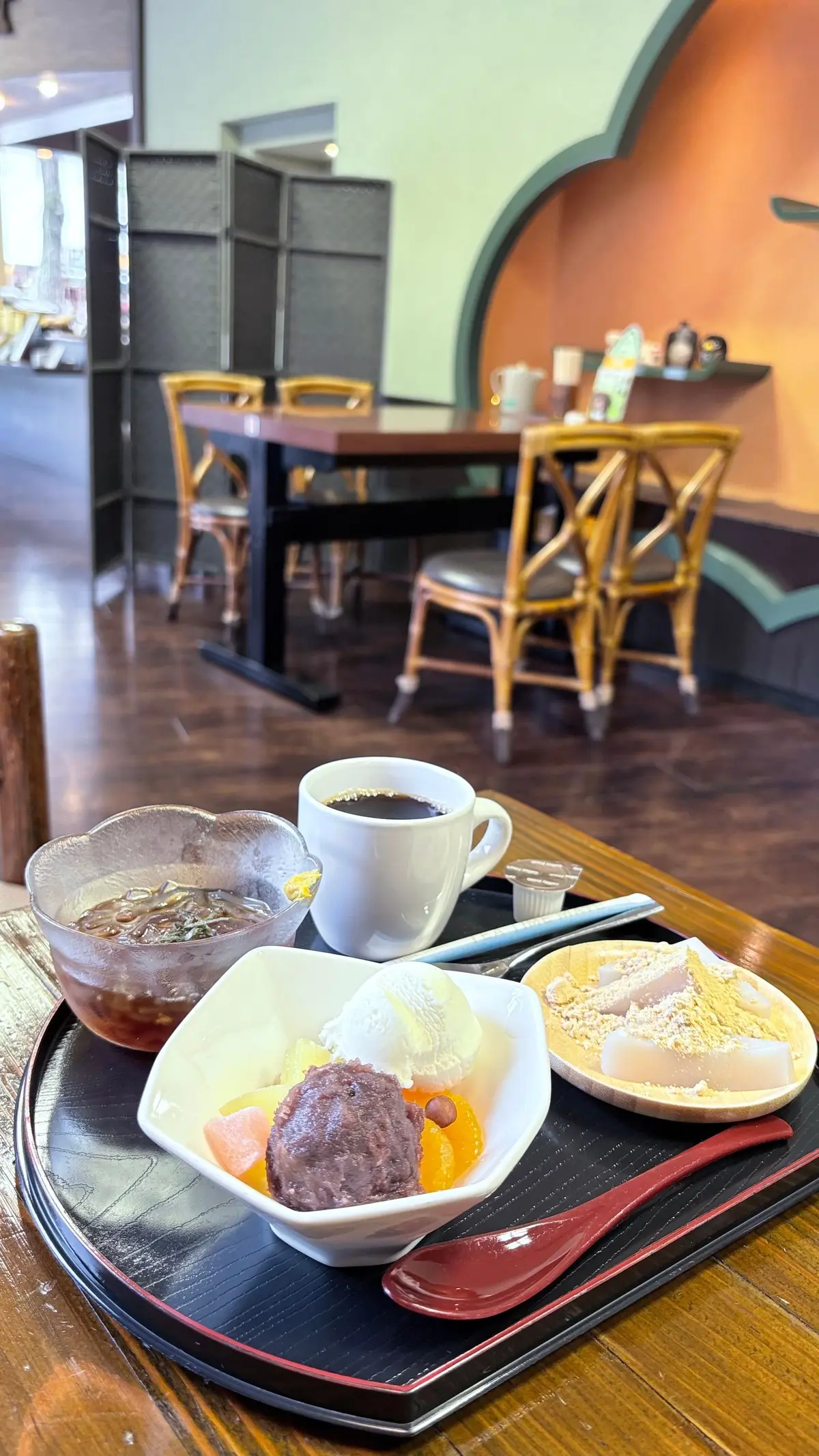 木製のトレイに、コーヒー、赤えんどう豆の甘味、白玉のデザートが置かれている。背景にはテーブルと椅子が見えるカフェの様子。