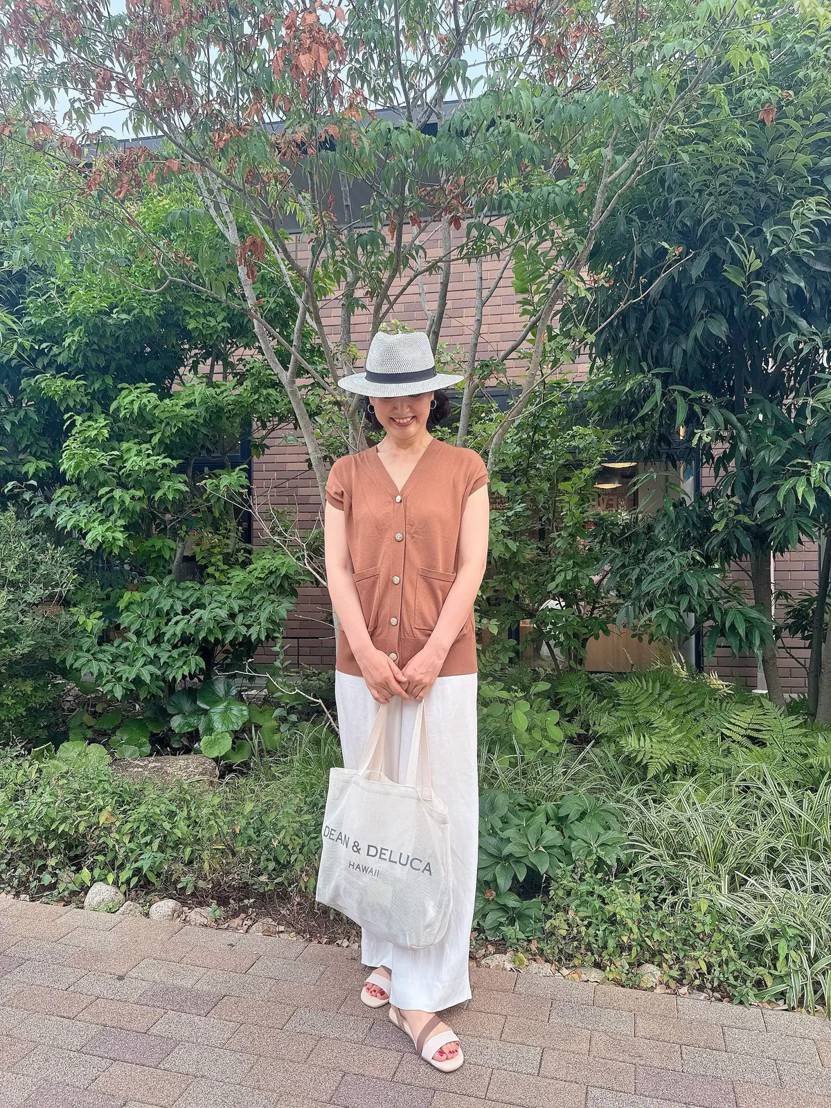 茶色のカーディガンに白いロングスカート、麦わら帽子を被った女性が笑顔で立っている。左手に「Dean & Deluca Hawaii」のロゴが入った白い紙袋を持つ。背景には緑豊かな木々が見える。