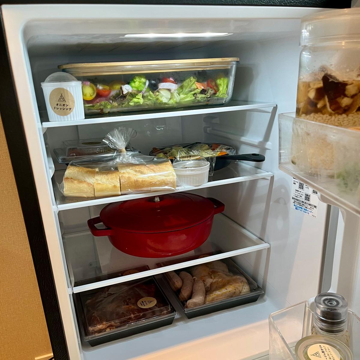冷蔵庫のドアが開いており、複数の棚に野菜、サンドイッチ、調理器具、肉類などが整理して並んでいる。