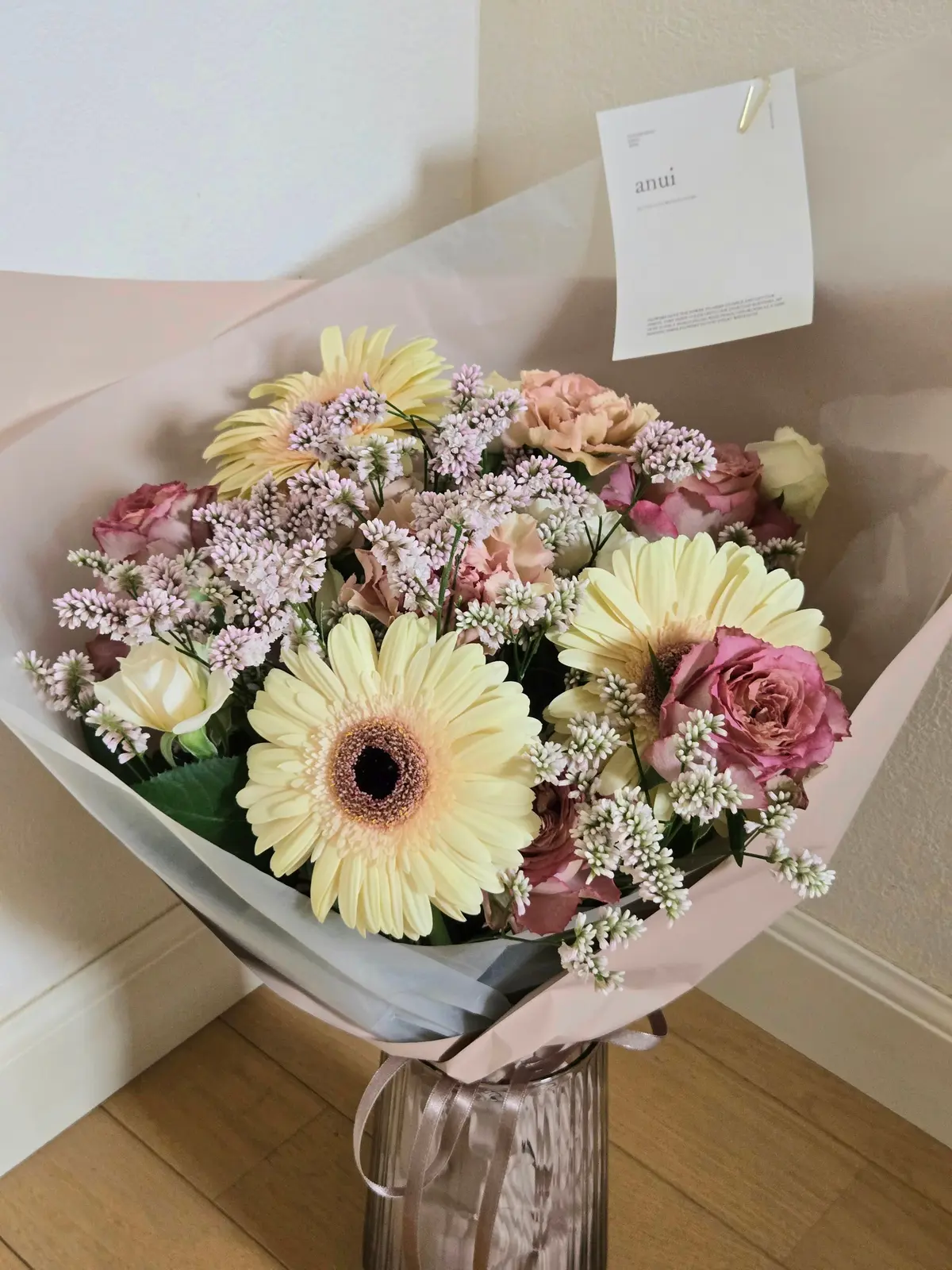 淡いピンクの紙で包まれた花束。黄色のガーベラ、ピンクのバラ、白い花、紫色の小さな花が混ざり、自然な配置。背景には白い壁と木の床。
