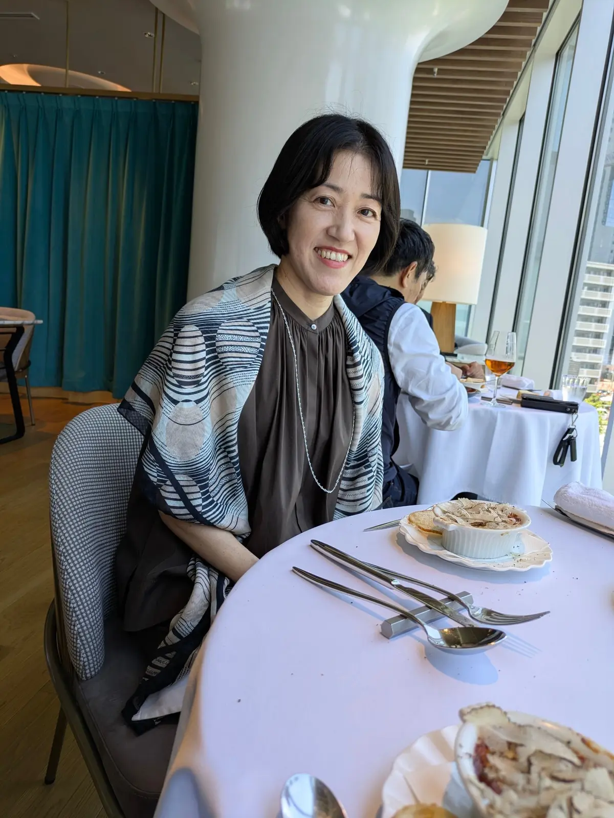 女性がレストランのテーブルに座り、料理と銀器が置かれた白いテーブルクロスをかけたテーブルに向かって微笑んでいる。背景には窓から街の景色が見え、青いカーテンと木製の天井が確認できる。
