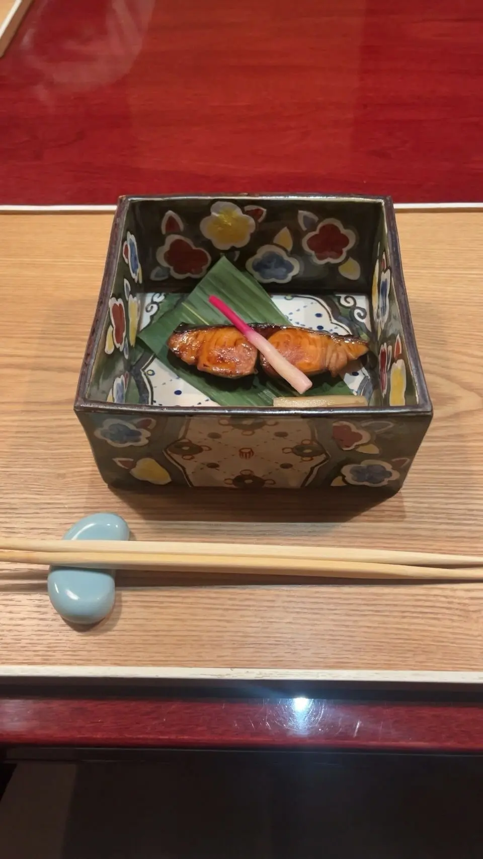 花模様の四角い器に、焼き魚と生姜の千切りがのせられた料理。木製のテーブルに、青い箸置と箸が置かれている。