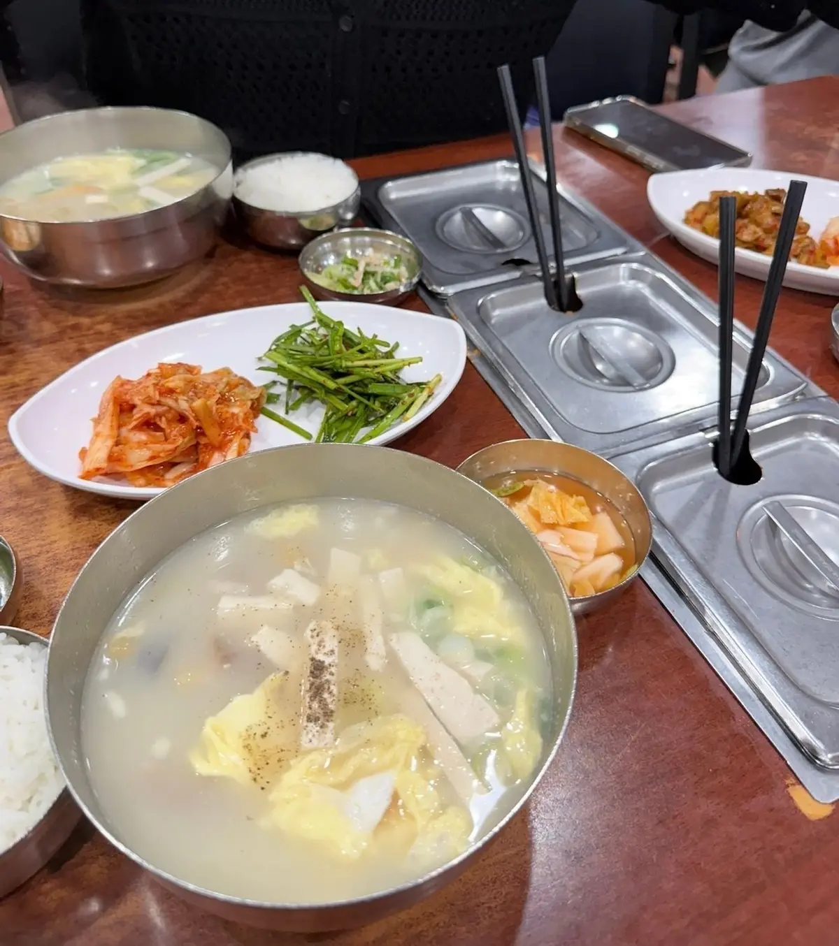 木製のテーブルに並べられた韓国料理。スープ、ご飯、キムチ、おかずの小鉢がステンレス製の容器に入っている。黒い箸が置かれている。