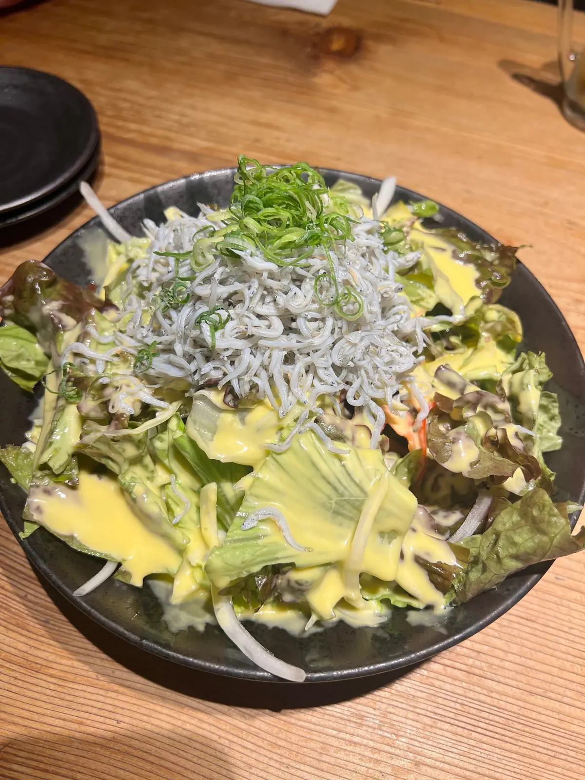じゃこサラダ。黒いお皿に盛り付けられた、レタスとアボカドのサラダ。黄色いドレッシングが全体にかけられ、上には細切りになったネギと海苔が散らされている。